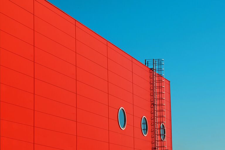 Red industrial rooftop wall with safety ladder ascending to sky against turquoise background