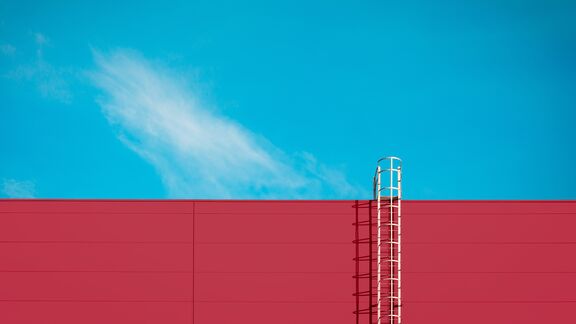Red industrial rooftop wall with white safety ladder ascending to sky against turquoise background